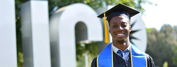 Photo of a student in a cap and gown in front of the UCR letter sculpture.